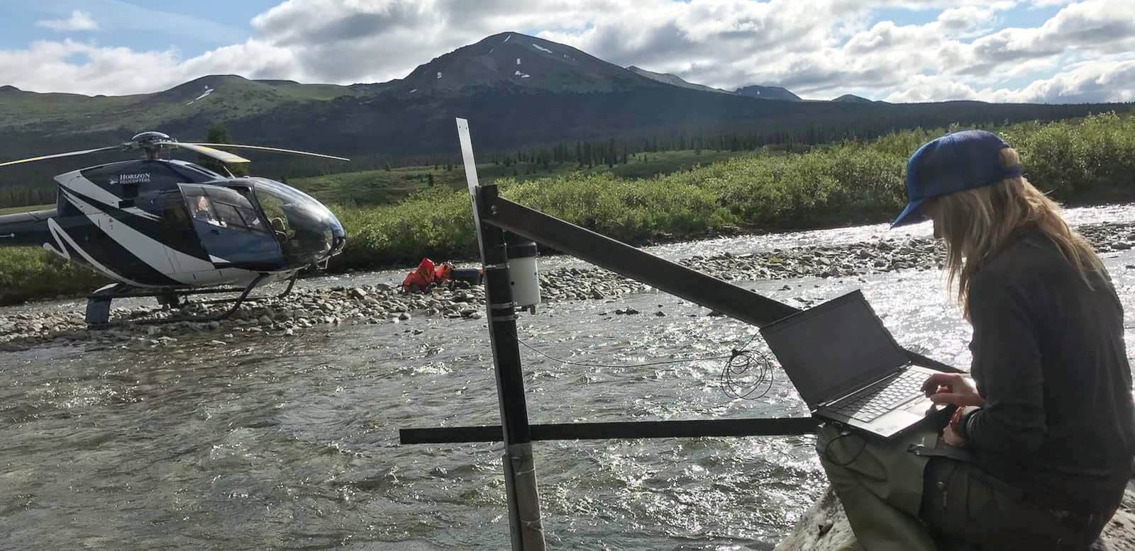 Crystal working one a laptop next to a stream with a helicopter on the other side and mountains in the near background